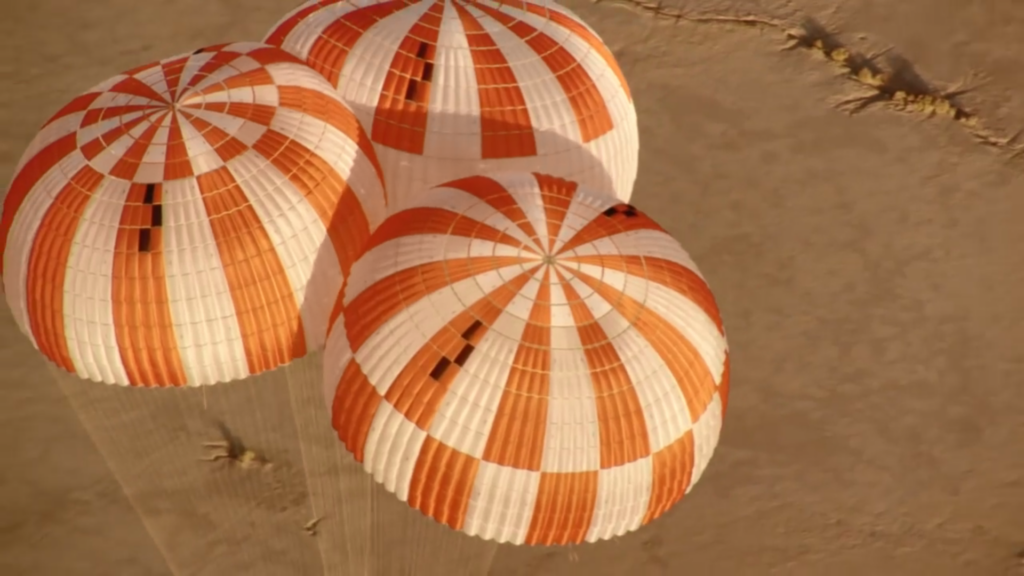 Three inflated red-and-white-striped parachutes drift over a cracked desert landscape.