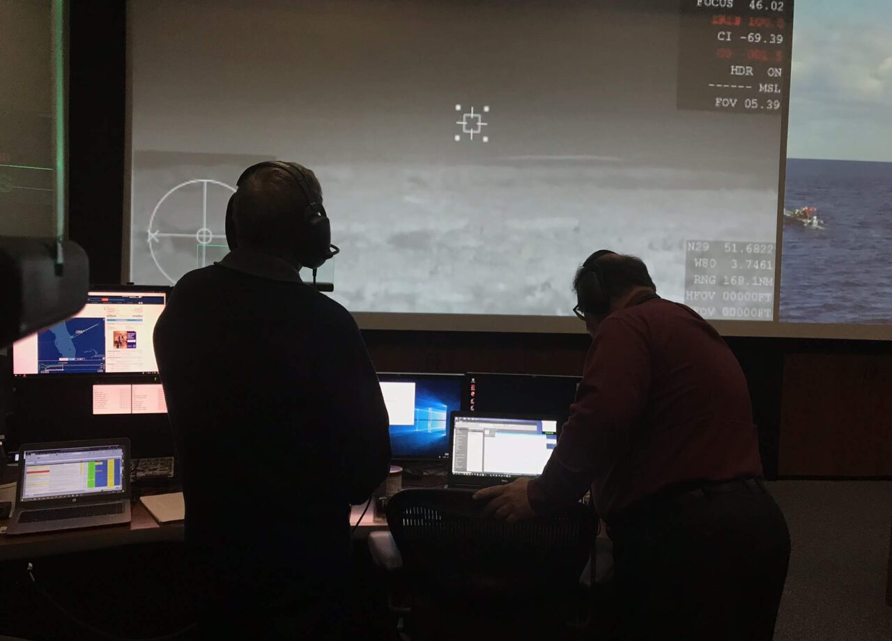 Rich Schwartz and Harry Verstynen are backlit by a screen displaying a cloudy sky in mission control
