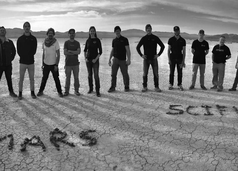 MARS and SCIFLI team members stand behind the acronyms "MARS" and "SCIFLI" spelled out with rocks on the cracked desert ground.