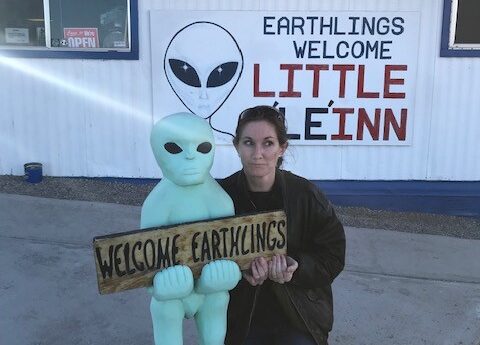 Jennifer Inman poses with a statue of a green alien. They hold a sign that says "Welcome Earthlings." A sign behind them on a building says "Earthlings Welcome: Little A'le'inn"