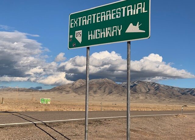 A sign says "Extraterrestrial Highway" in front of a deserted two-lane highway in the desert. There is a SCIFLI sticker on the sign, and another sign with a green alien on it in the distance.